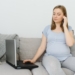 Attractive pregnant woman touching tummy and using laptop on bed