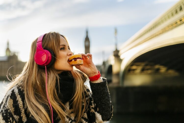 UK, London, young woman listening music and taking a snack near Westminster Bridge